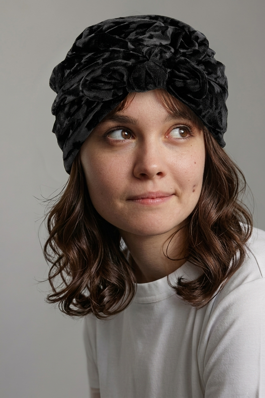 A woman wearing a black textured fabric headband with large ruffled flowers, looking to the side against a plain light gray background.