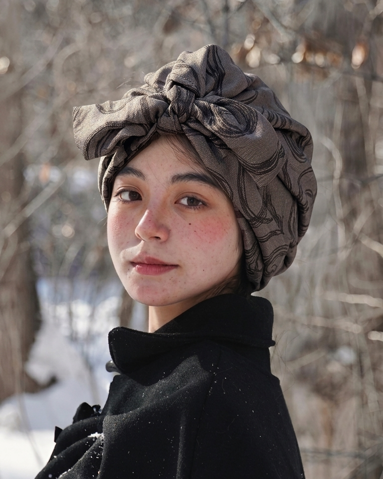 A woman outdoors in a snowy setting wearing a dark patterned fabric headwrap turban and a black coat.