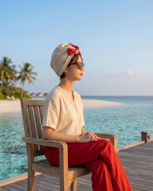 A woman sits on a wooden chair on a tropical beach boardwalk, wearing a cream linen top, bright red pants, a beige head wrap with a red flower, and sunglasses, looking out toward the ocean.